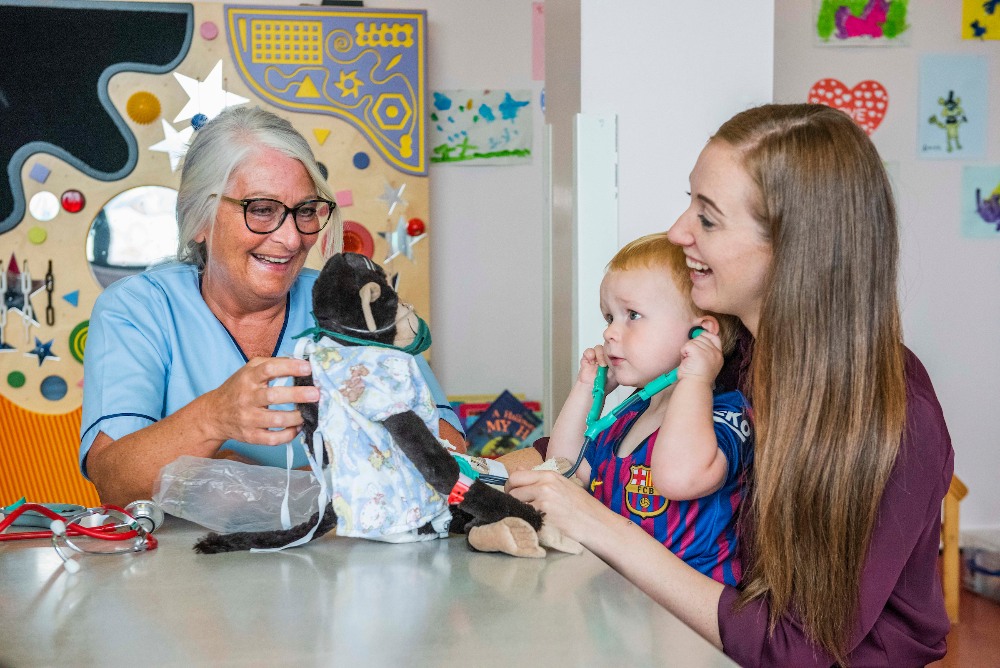 There is a play specialist holding a toy monkey. Next to the play specialist there is a woman who is smiling and holding a little boy in football uniform. He is using Stethoscope on a toy monkey
