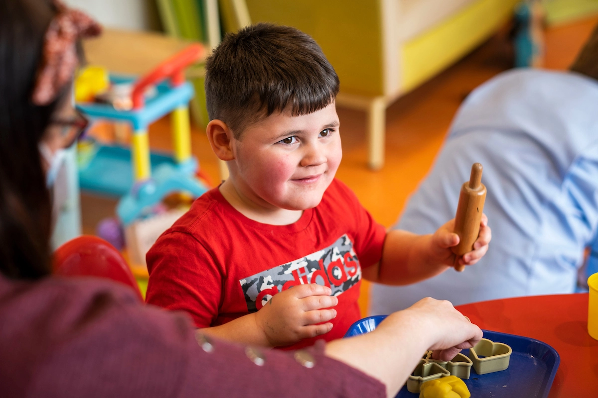 A child, wearing a red t-shirt, is holding a toy and smiling.