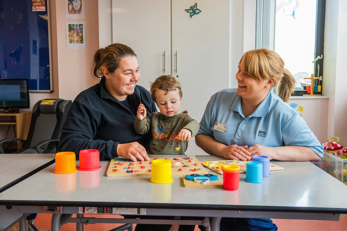 2 women and a toddler are sitting at the table. A woman on the right is wearing a black jumper and smiling. She is holding a child. A woman on the left is wearing blue uniform and is also smiling. 