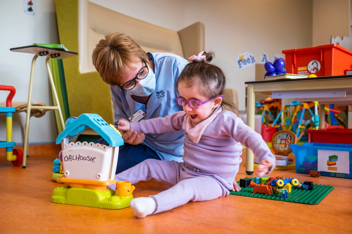 A little girl, wearing purple pyjamas, is playing in the playroom and smiling. A play specialist in a blue uniform, glasses and face mask is looking at her.