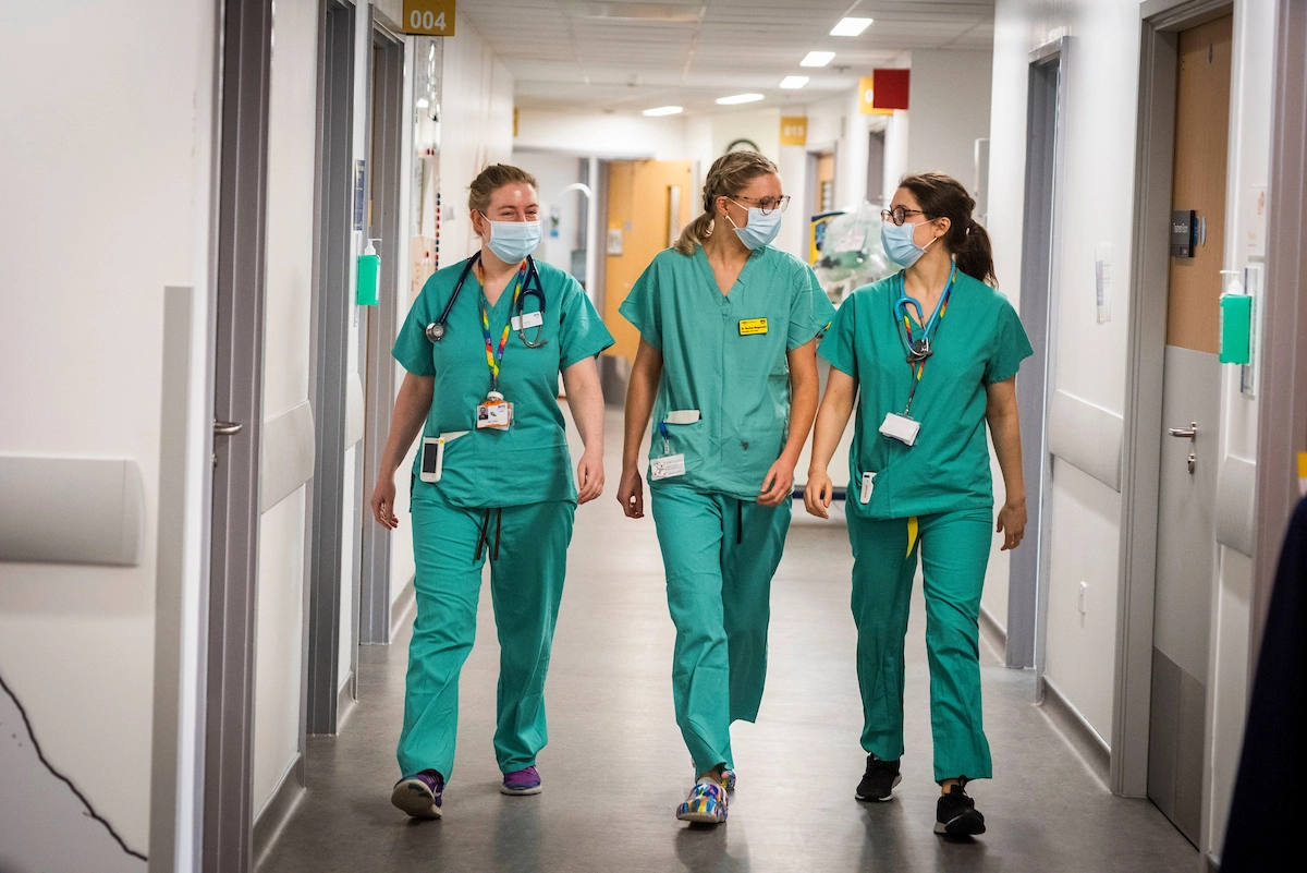 3 female nurses, wearing a green unifrom, are walking in the hall and smiling
