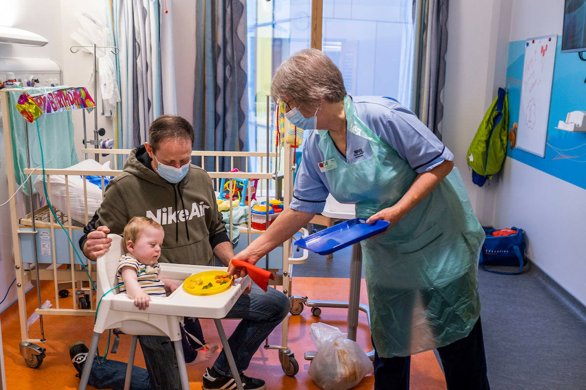 A father, in a green hoodie and jeans, is sitting next to his baby daughter. His baby daughter is wearing a striped t-shirt and is sitting in a baby chair. A nurse is giving her food