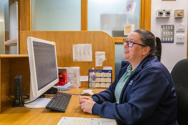 a woman in a blue navy jacket in glasses sitting in front of a laptop at the reception desk and smiling