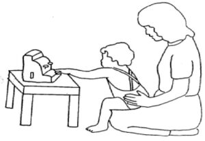 An adult is Kneeling on the floor in front of a coffee table. A child is sitting on adult's lap, with their hips and knees at 90 degrees and their feet are flat on the floor. The child is trying to reach a toy placed on the table 