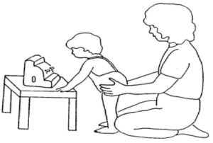 The child has straightened their knees and trying to reach a toy on a table while an adult is supporting them around their hips