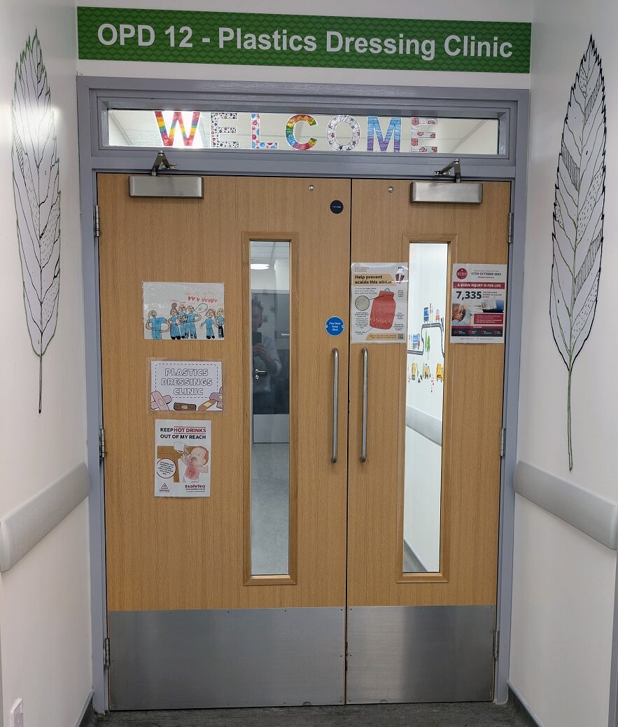 The image shows the entrance to the OPD 12 - Plastics Dressing Clinic. The double wooden doors have small vertical glass panels and are decorated with posters and notices. The walls feature simple feather illustrations.