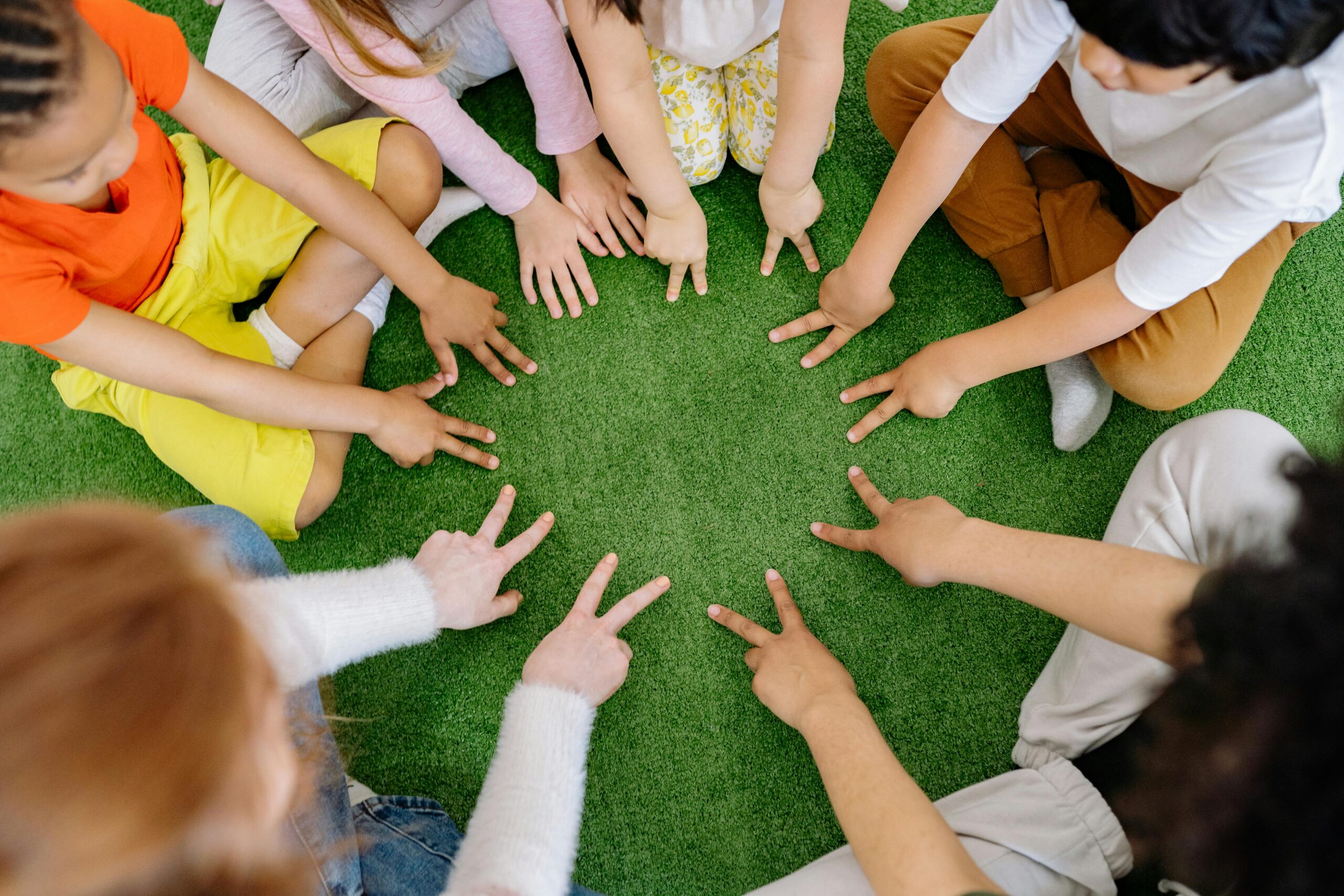A group of children sitting in a circle on artificial green grass, extending their hands towards the center while making playful gestures with their fingers. The image captures a top-down view