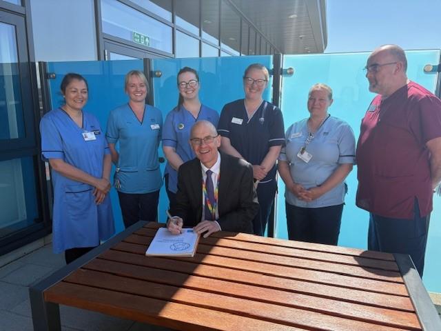 A group of healthcare staff stand around a man in a suit who is seated at an outdoor table signing a document. The group is smiling and posing together outside a hospital building on a sunny day.