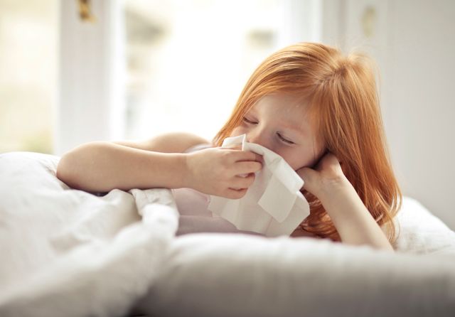 Redheaded child resting in bed, wiping nose with tissue, unwell with flu-like symptoms